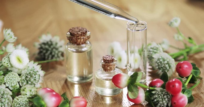 Production of homemade perfume concept. Perfumer makes aroma liquids parfume poures essential oil pipette to glass vial and closes cork cap, hand closeup. Fresh flowers near bottles on wooden table.