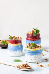 Vegan blue spirulina chia puddings with jam and granola in glass jars on a light blue table. Concept of healthy eating, dieting, fitness menu. Various layered dessert for breakfast. Top view.