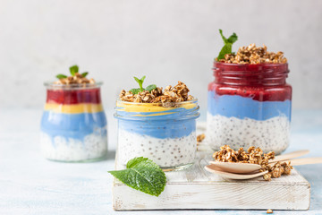 Vegan blue spirulina chia puddings with jam and granola in glass jars on a light blue table. Concept of healthy eating, dieting, fitness menu. Various layered dessert for breakfast. Top view.