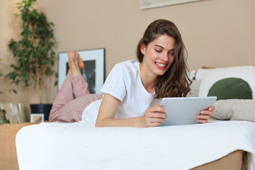 Smiling woman is liying on the bed and use her digital tablet.