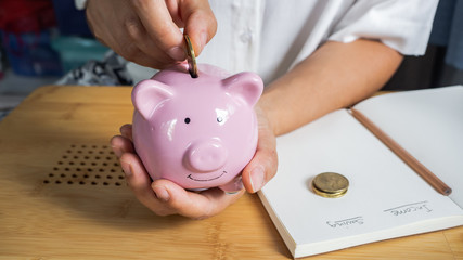 A hand is putting the coins in a pink piggy bank isolate in wooden background.