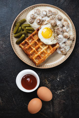 Belgian waffle with champignons, fried egg and pickles, flatlay on a dark brown stone background, vertical shot