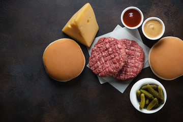 Raw beef burger cutlets and burger buns over dark brown metal background, horizontal shot with space, top view