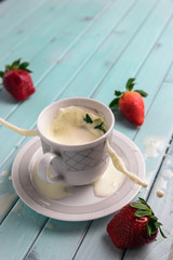 Red ripe strawberries and a white coffee Cup, on a turquoise wooden table. The berry falls into the milk. Frozen movement. Splashes from the Cup.