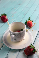 Red ripe strawberries fall into a Cup of milk. A Cup of milk on a turquoise wooden table. Splashes of milk.