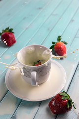 Red ripe strawberries and a white coffee Cup, on a turquoise wooden table. The berry falls into the milk. Frozen movement. Splashes from the Cup.