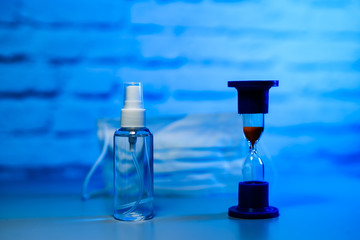 Protective mask and antiseptic on a blue background. Sand watch as a symbol of time. Sanitizer, protective face mask. Hygiene concept.