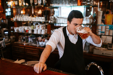 A young caucasian  waiter drinks beer at a bar