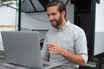 A man wearing a long sleeve grey button down business shirt with brown slicked comb over hair working on laptop on a picnic table remotely near his camper able to avoid the busy society work community