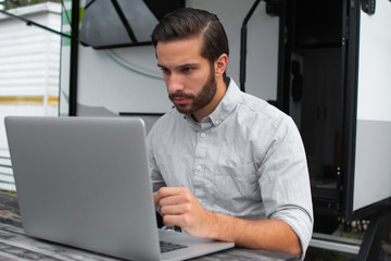 A man wearing a long sleeve grey button down business shirt with brown slicked comb over hair working on laptop on a picnic table remotely near his camper able to avoid the busy society work community