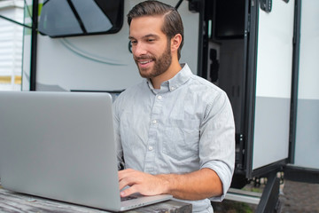 A man wearing a long sleeve grey button down business shirt with brown slicked comb over hair working on laptop on a picnic table remotely near his camper able to avoid the busy society work community