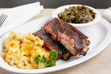 A view of a plate of barbecue spare ribs, with a side of macaroni and cheese, as well as collard greens, in a restaurant or kitchen setting.
