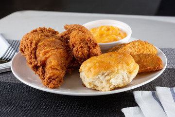 A view of a plate of fried chicken, with a side of biscuit, potato wedges and cup of macaroni and cheese, in a restaurant or kitchen setting.