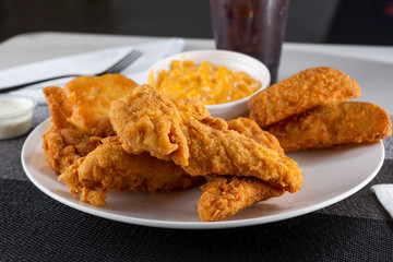 A view of a plate of breaded chicken tenders, with potato wedges and macaroni and cheese sides, in a restaurant or kitchen setting.