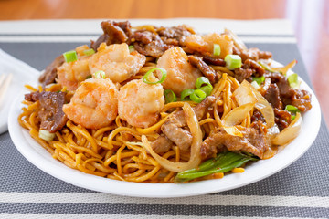 A view of a plate of Chinese chow mein featuring shrimp, beef and chicken, in a restaurant or kitchen setting.