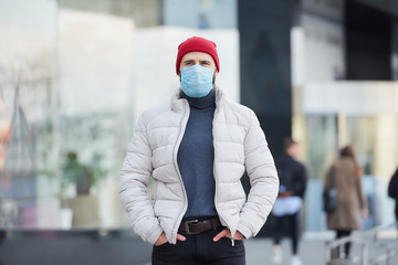 Fototapeta premium A man wearing a medical face mask to avoid the spread coronavirus (COVID-19). A guy with a mask on the face because of the pandemic holding hands in the pockets of the jeans in the center of the city.