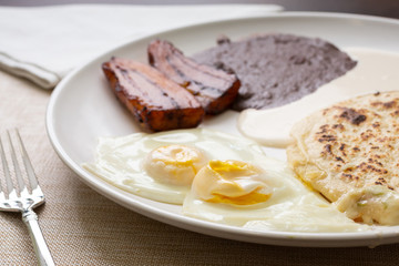 A view of a Latin American breakfast plate in a restaurant or kitchen setting, featuring sunny side up eggs, fried plantains, pupusas and black beans.