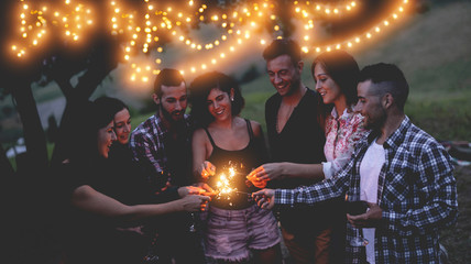 Happy friends having fun at bbq dinner with sparklers lights outdoor while drinking wine - Young...