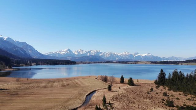 Hoch &uuml;ber dem Bannwaldsee mit Blick auf die Tannheimer Berge mit schneebedeckten Gipfeln im Fr&uuml;hjahr mit Buchinger Bach im Vordergrund