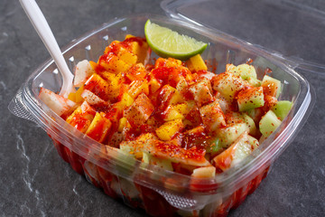 A view of a plastic container of Mexican fruit salad, featuring lime chili seasoning and Chamoy sauce, in a restaurant or kitchen setting.