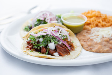 A view of two tacos al pastor with a side of beans and rice, in a restaurant or kitchen setting.