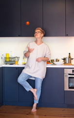 A young woman with short hair juggles a tomato standing in her blue kitchen