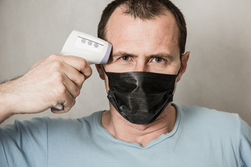 A depressed man wearing protective mask ready to use infrared forehead thermometer to check body temperature for virus symptoms - epidemic virus outbreak concept. Coronavirus.Thermometer gun