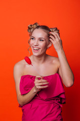 The girl in a red dress on a orange background in the studio. Blonde girl with two hair knots holding red heart on the stick, looking to the camera and smiling.