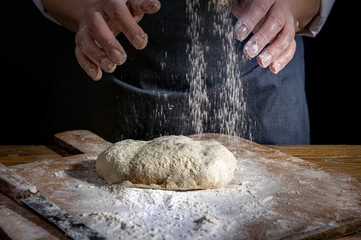 Female hands knead the dough on a wooden antique table on a dark background, close-up, shallow depth of field, beautiful directional lighting. Concept of home baking and comfort.