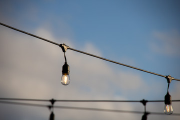 Obraz premium A string of outdoor lighting with on and off light bulbs hanging in the air, against a bright blue sky with clouds. Early morning feeling, selective focus on light bulbs.