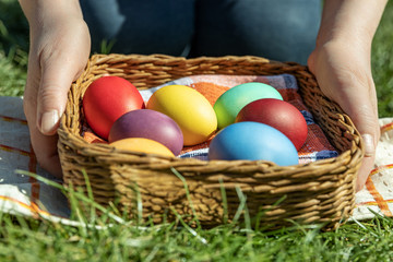 Fototapeta na wymiar Female hands hold multi-colored Easter eggs in a basket on the grass, the background is blurred, shallow depth of field, selective focus. Easter holiday concept
