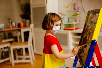 Portrait of a small preschool child with virus protection surgical face mask due to coronavirus, painting at an art easel at home