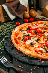 Delicious pizza with mushrooms, olives and pepper on the black board on the dark wooden table with herbs and tomatoes, vertical, side view
