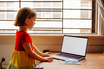 Portrait of a small preschool child with laptop computer and surgical face mask, using internet online at home by the window
