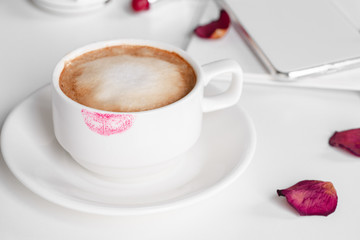 Lipstick mark on cup of cappuccino on white background with rose petals. Morning romance concept.