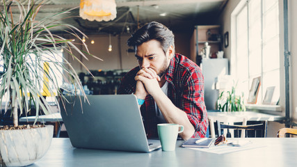 Young man working with laptop. Entrepreneur wearing casual clothes and stubble in big spacious bright room. Workspace on the table