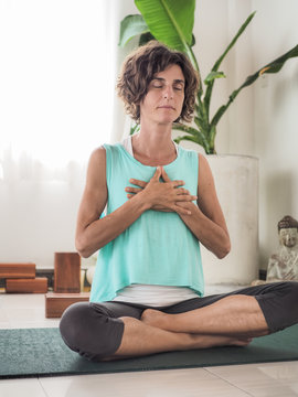 Meditating Woman With Eyes Closed And Hands In Heart Mudra In Crossed Legged Lotus Position On A Yoga Mat In A Studio With Cushions Buddha And Palm Tree