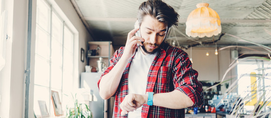 Young man working. Entrepreneur wearing casual clothes and stubble in big spacious bright room. Workspace on the table. Talking on mobile phone