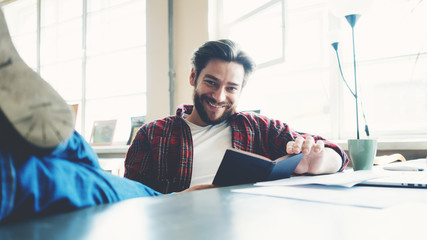 Smiling man with stubble and casual clothes reading book while sitting in big spacious working room