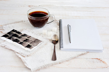 A cup of coffee, chocolate, a sheet of paper and a pen on a white wooden surface. Background for...