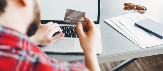 Man using credit bank card to pay online with laptop. Wearing red casual shirt. Close-up. Wide screen