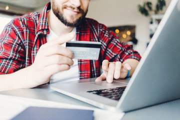 Young man using credit bank card to pay online with laptop. Wearing red casual shirt and sitting in...