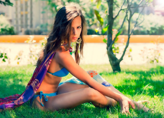 An slim young girl in a blue bikini and an blue scarf in her neck sitting on the grass of a...