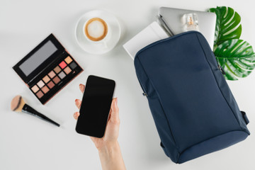 Female hand holding a smartphone with mockup. Flat lay of backpack with digital devices, coffee and cosmetics on white desk. Freelance and telework concept