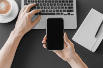 Male hand holding a smartphone with mockup on workspace background with coffee and laptop. Online working concept.
