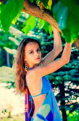 Portrait of beautiful young girl in blue swimsuit and long earrings with dark eyes and long hair posing near a tree on river bank.