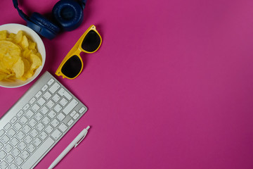 Flat lay of keyboard, headphones, sunglasses, pen, and a plate of chips in purple background....