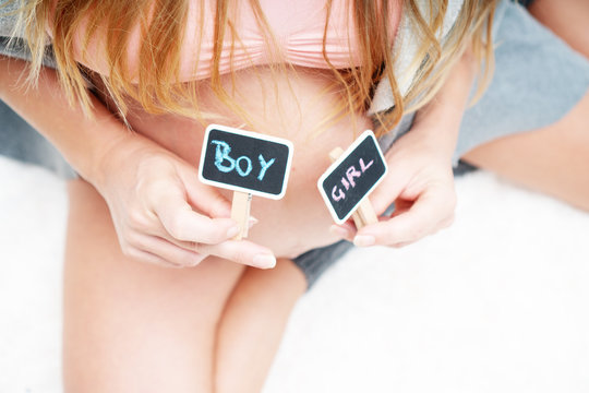 Cropped image of pregnant woman holding mini blackboards with written boy and girl while sitting on her bed at home. Maternity, pregnancy, new life and family planning concept