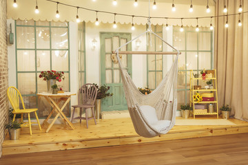 Suspended wicker swing on the summer porch with garlands of light bulbs. Swing hammock made of thread on the background of the house verandah. Terrace of the summer house design of the photo Studio.
