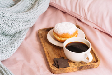 Morning breakfast in bed with fresh and hot bracing coffee, a doughnut in powdered sugar and three slices of chocolate on a wooden tray against a backdrop of pink sheets and blue plaid